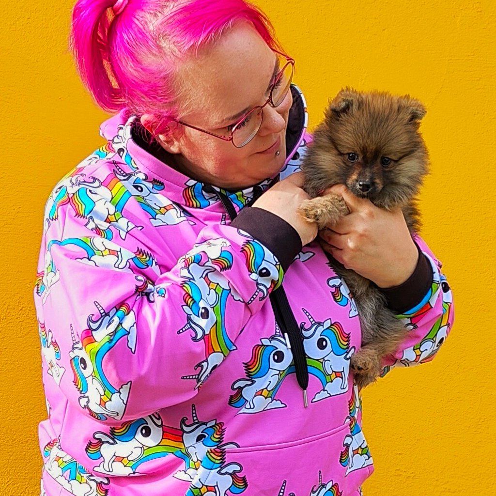 A lady holding her dog and wearing a SHALMIAK hoodie with colorful unicorns on pink background.